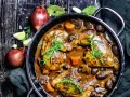 Herzhaftes Coq au Vin in einem gusseisernen Topf, mit Pilzen, Karotten und Kräutern garniert. Daneben Zwiebeln und Knoblauch.