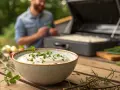 Schüssel mit cremiger Sour Cream, garniert mit Petersilie, steht auf einem Holztisch. Im Hintergrund bereitet ein Mann das Grillgut vor.