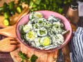 Omas Gurkensalat mit Joghurt, frisch zubereitet und mit Kräutern verfeinert, serviert in einer rosa Schüssel auf einem Holzbrett.