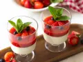 Zwei Gläser mit cremiger Panna cotta, garniert mit Erdbeersauce, frischen Erdbeeren, Blaubeeren und Minze.