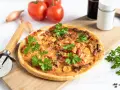 Flammkuchen mit Pfifferlingen, garniert mit Petersilie, auf einem Holzbrett. Tomaten und Schalotten im Hintergrund.