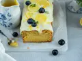Ein saftiger zitronen schmand kuchen, dekoriert mit Zitronenscheiben, Blaubeeren und Zitrusschale. Daneben eine Kanne mit Zitronensauce.