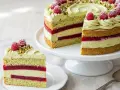 Eine wunderschöne Pistazien Himbeer Torte mit frischen Himbeeren und gehackten Pistazien. Ein Stück ist bereits auf einem Teller angerichtet.
