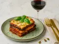 Schicht für Schicht ein Genuss: Ein Stück authentisches Melanzane Rezept Original, garniert mit frischem Basilikum, neben einem Glas Rotwein.