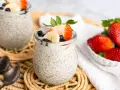 Zwei Gläser mit cremigem Chia Pudding, garniert mit Erdbeeren, Bananen und Blaubeeren. Daneben eine Schale voller frischer Erdbeeren.