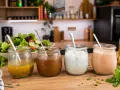 Vier Gläser mit verschiedenen **Salatdressing**-Sorten stehen auf einem Holztisch. Im Hintergrund ein frischer Salat.