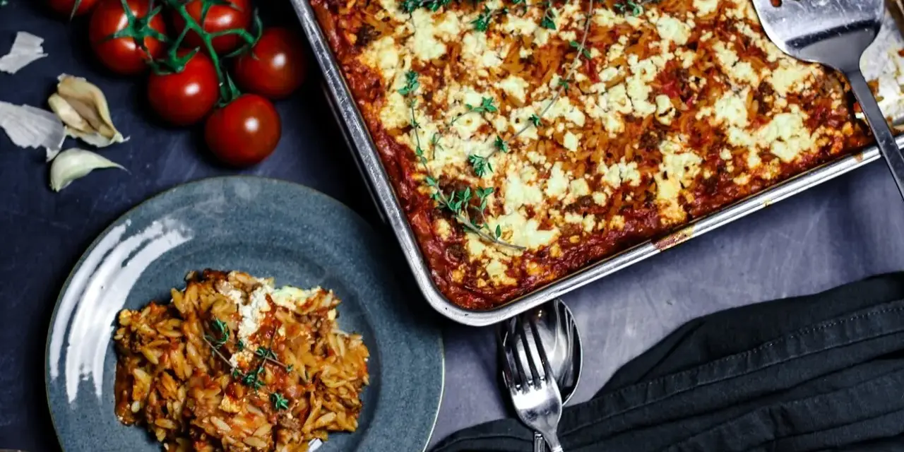 Herzhafter Kritharaki Auflauf mit Hackfleisch und Feta, garniert mit Thymian. Daneben frische Tomaten und Knoblauch.
