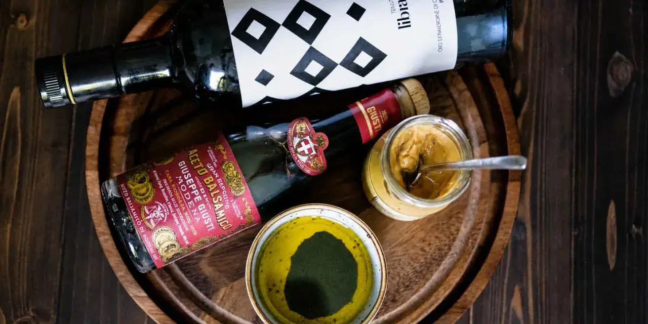 Zutaten für ein köstliches Balsamico-Dressing: eine Flasche Olivenöl, eine Flasche Aceto Balsamico Giuseppe Giusti und eine Schale mit grünen Kräutern.