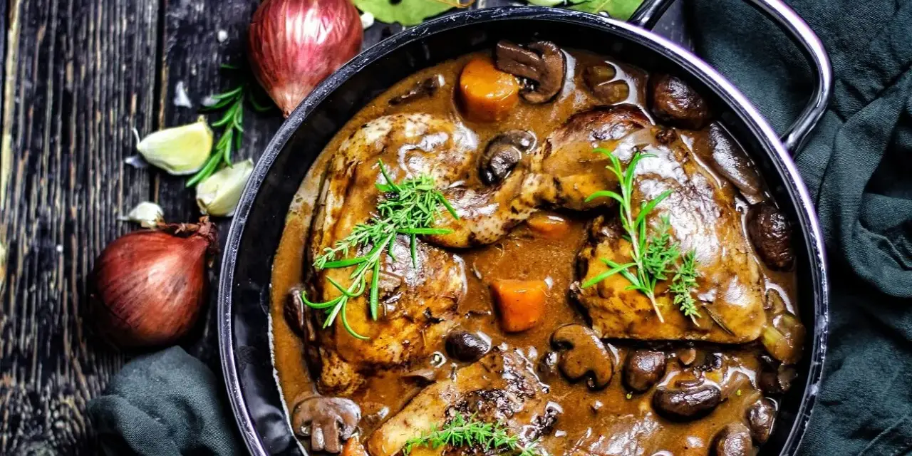 Herzhaftes Coq au Vin in einem gusseisernen Topf, mit Pilzen, Karotten und Kräutern garniert. Daneben Zwiebeln und Knoblauch.