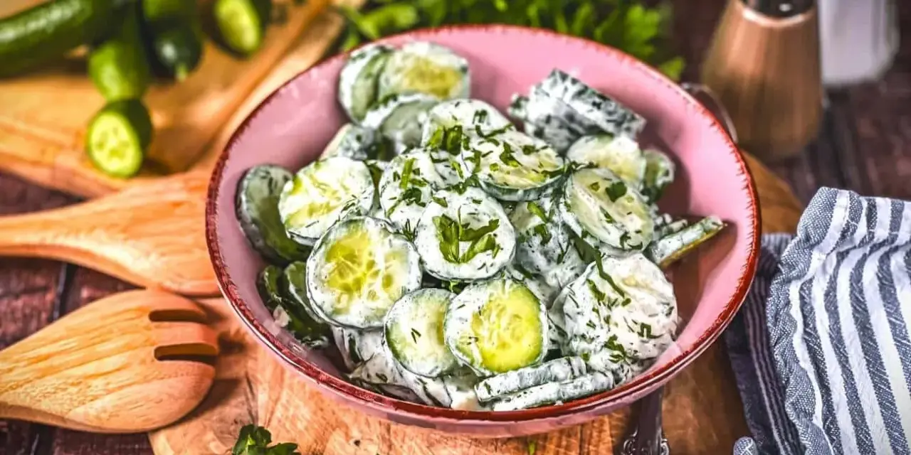 Omas Gurkensalat mit Joghurt, frisch zubereitet und mit Kräutern verfeinert, serviert in einer rosa Schüssel auf einem Holzbrett.