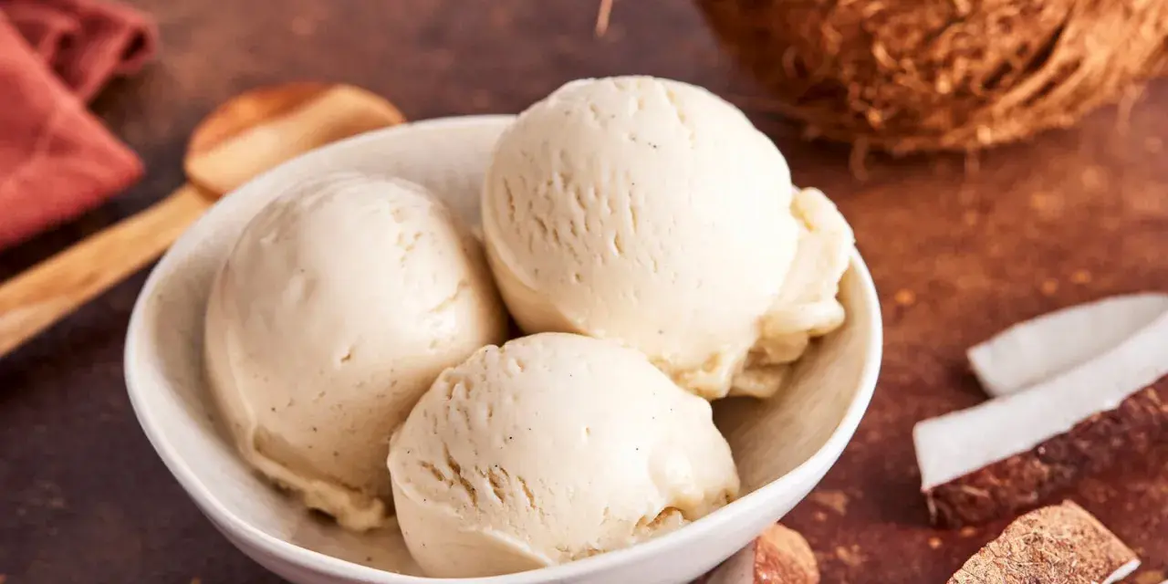 Schaufeln cremiges Kokoseis, perfekt zum Kokoseis selber machen mit Eismaschine. Daneben Kokosnussstücke.
