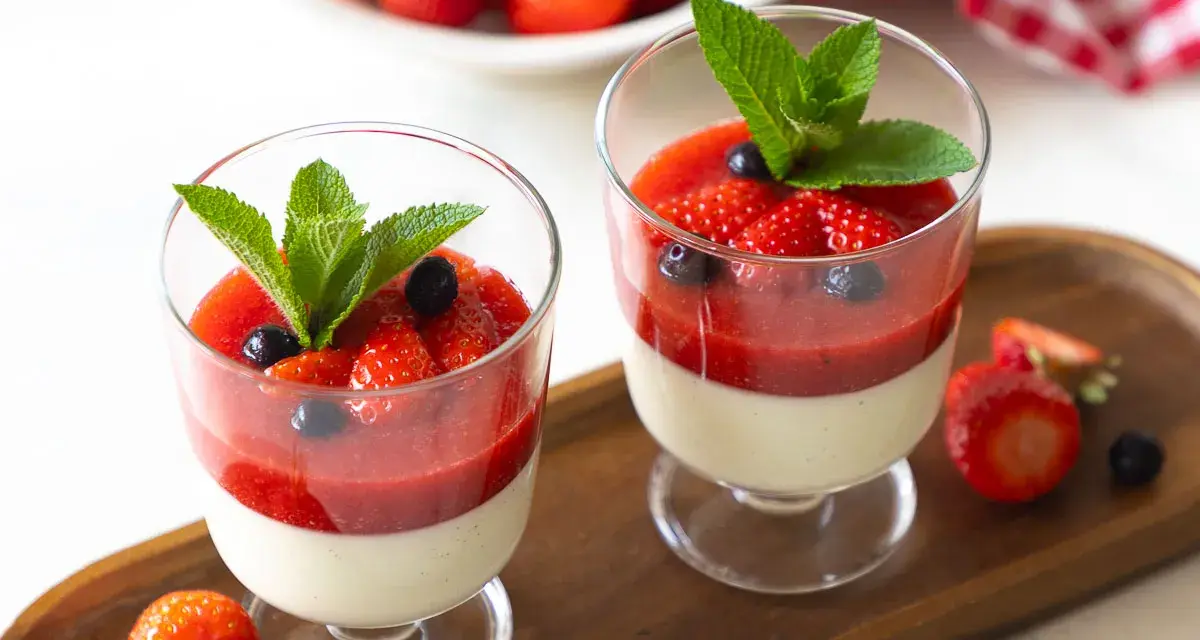 Zwei Gläser mit cremiger Panna cotta, garniert mit Erdbeersauce, frischen Erdbeeren, Blaubeeren und Minze.