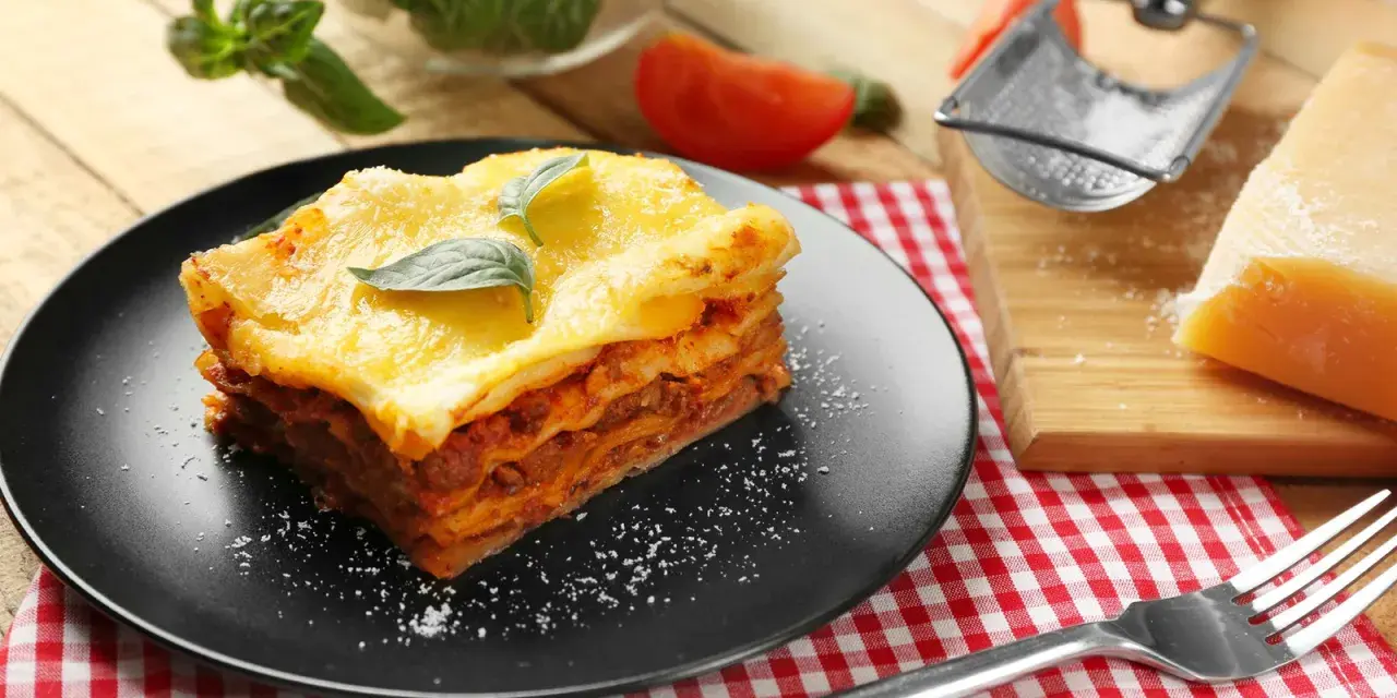 Ein Stück Lasagne mit Hackfleischsauce und cremiger Bechamelsoße, garniert mit Basilikum. Daneben ein Stück Parmesan und eine Gabel.