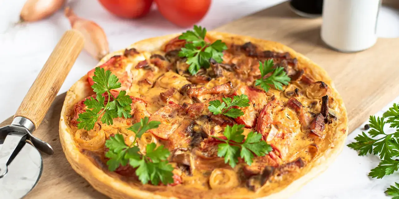 Flammkuchen mit Pfifferlingen, garniert mit Petersilie, auf einem Holzbrett. Tomaten und Schalotten im Hintergrund.
