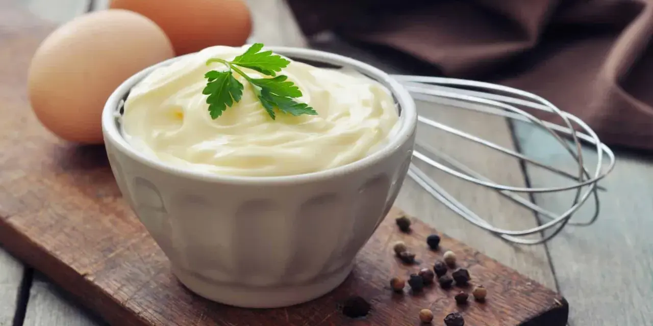 Cremige Mayonnaise in einer Schüssel, garniert mit Petersilie. Eier und ein Schneebesen deuten auf ein hausgemachtes mayonnaise rezept hin.