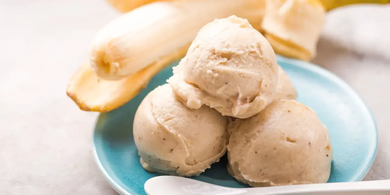 Cremiges Bananeneis, ganz einfach eis selber machen ohne eismaschine. Dazu reife Bananen und eine weiße Löffel auf blauem Teller.