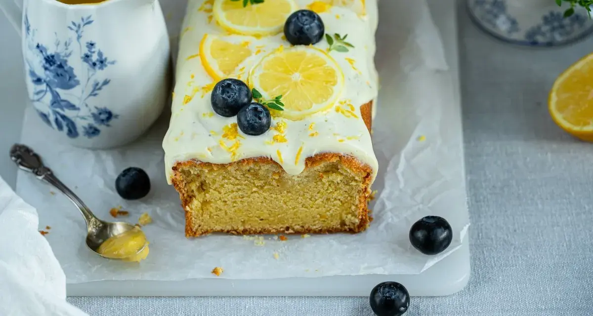 Ein saftiger zitronen schmand kuchen, dekoriert mit Zitronenscheiben, Blaubeeren und Zitrusschale. Daneben eine Kanne mit Zitronensauce.