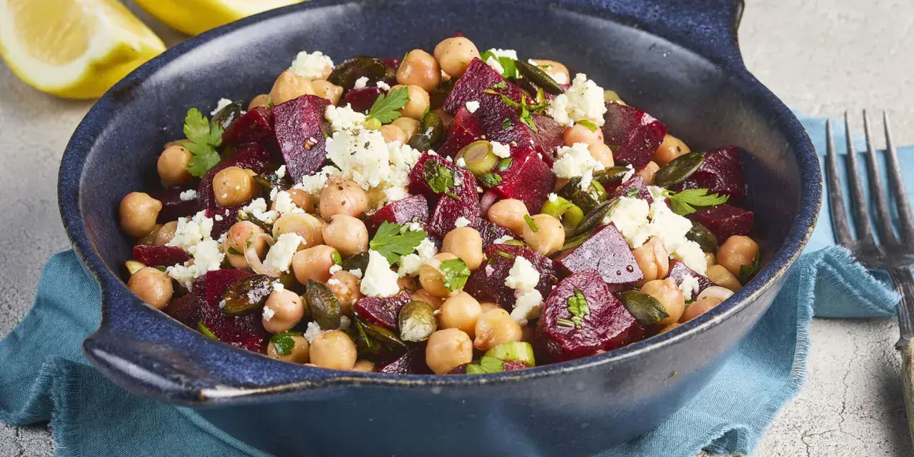 Ein bunter rote Bete Kichererbsen Salat mit Feta und Kürbiskernen in einer blauen Schüssel, garniert mit Petersilie und Zitronenspalten.