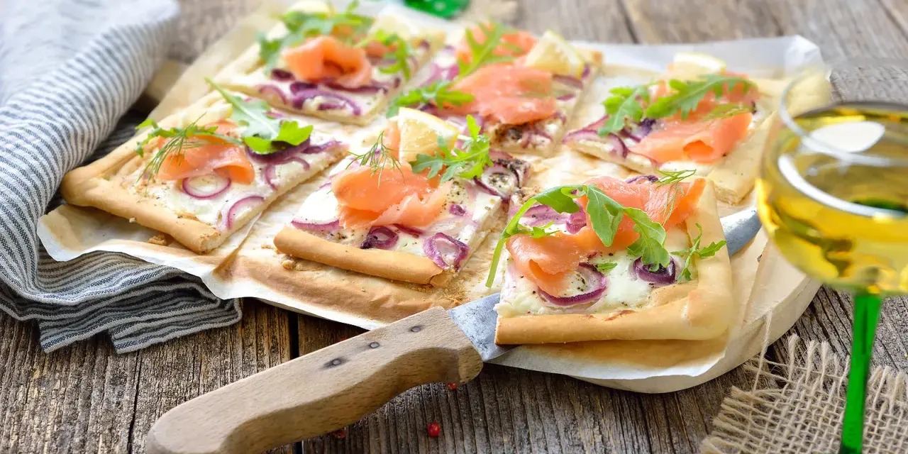 Flammkuchen mit Lachs und Crème fraîche, garniert mit Rucola und Dill, auf rustikalem Holztisch.
