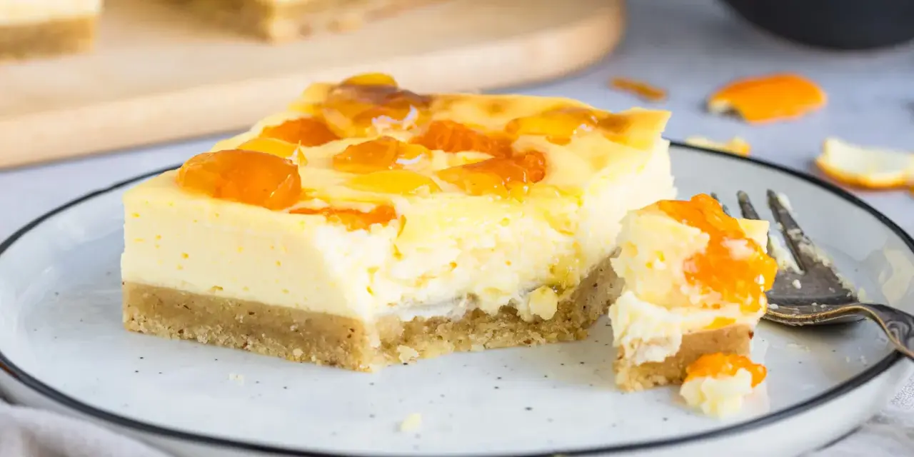 Ein Stück Mandarinen Schmand Kuchen Blech mit fruchtiger Glasur auf einem Teller, daneben ein Stück mit einer Gabel.