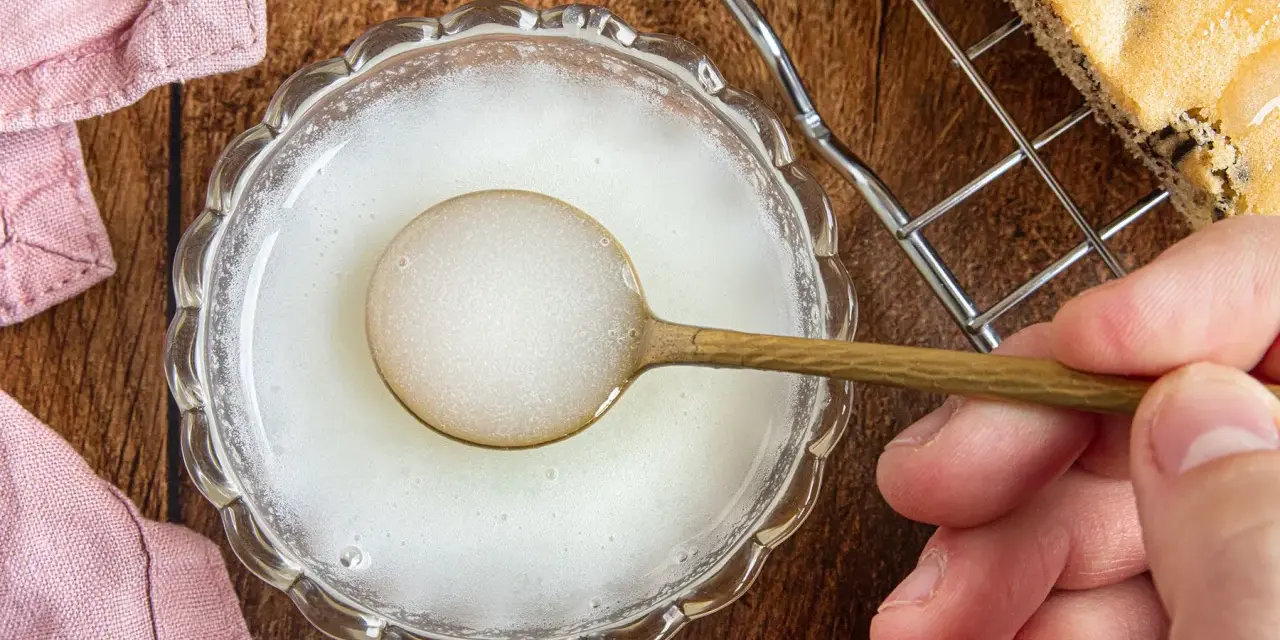 Ein Löffel mit zuckerguss ohne puderzucker in einer Schale. Daneben ein Stück Kuchen auf einem Gitter.