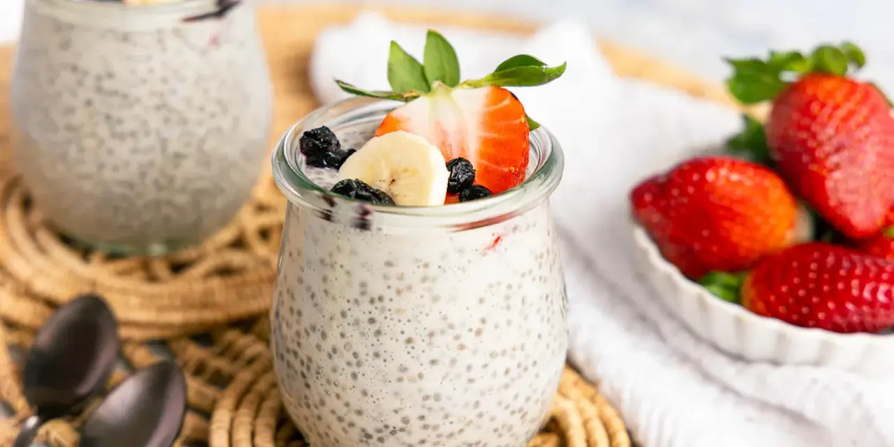 Zwei Gläser mit cremigem Chia Pudding, garniert mit Erdbeeren, Bananen und Blaubeeren. Daneben eine Schale voller frischer Erdbeeren.