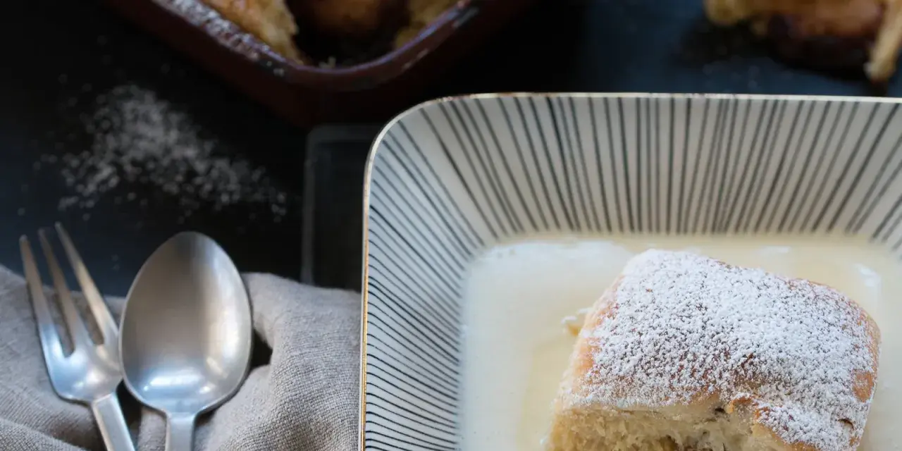 Buchteln original rezept: Ein Buchtel in Vanillesauce, bestäubt mit Puderzucker. Im Hintergrund weitere Buchteln im Ofen.