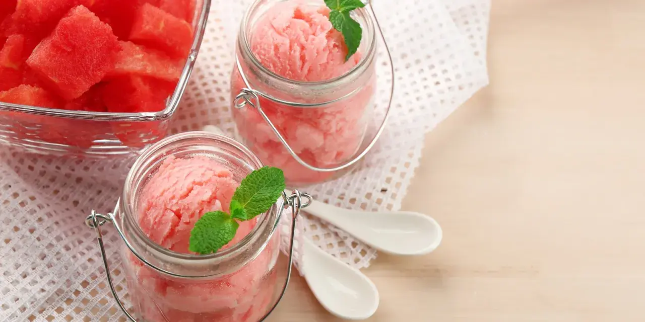 Erfrischendes Meloneneis selber machen: Rosa Sorbet in Gläsern mit Minze, daneben eine Schale mit Wassermelonenwürfeln.