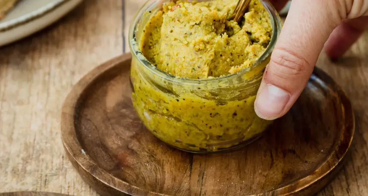 Ein Glas mit selbstgemachter Pistaziencreme, die mit einem goldenen Löffel umgerührt wird. Perfekt zum Brot oder als Dip.