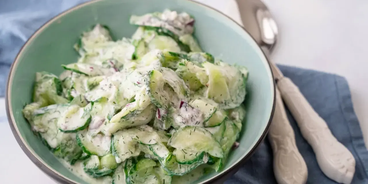 Frischer gurkensalat mit saurer sahne, garniert mit Dill und roten Zwiebeln, serviert in einer hellgrünen Schüssel.