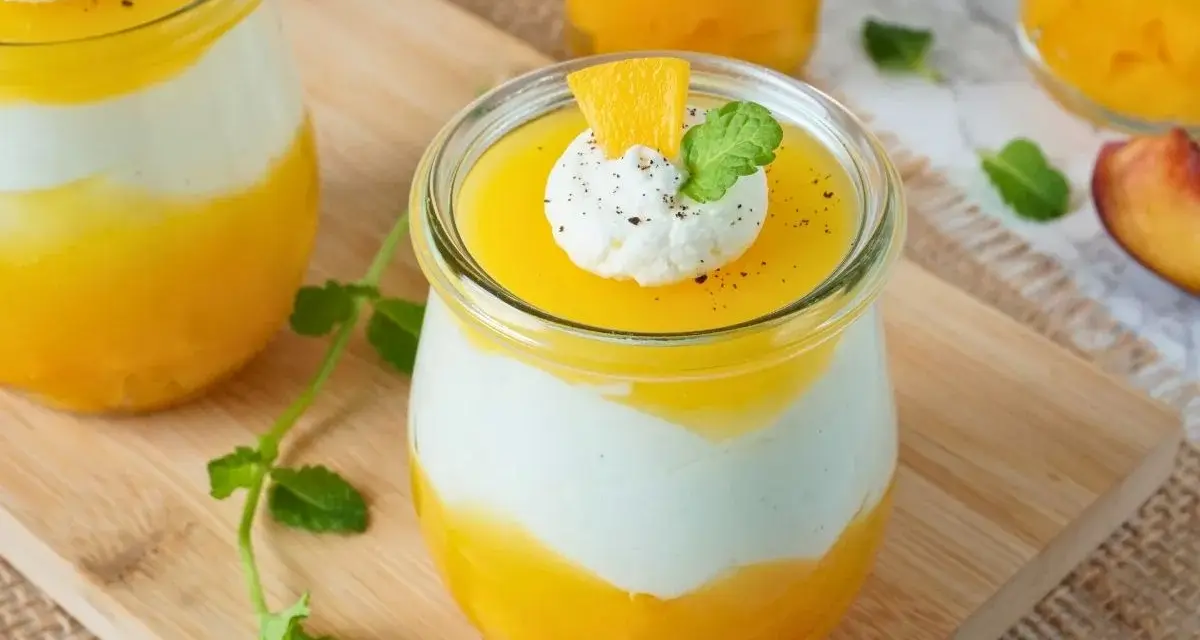 Erfrischendes Solero Dessert im Glas mit Mango-Fruchtsoße, weißer Creme und einem Klecks Sahne, garniert mit Minze und Orangenspalte.