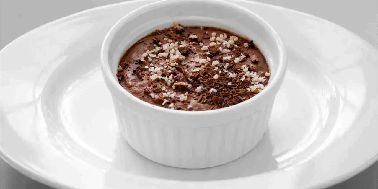 Ein cremiges Mousse au Chocolat, garniert mit Schokoladenraspeln und feinen Salzkristallen, serviert in einer weißen Schale auf einem Teller.