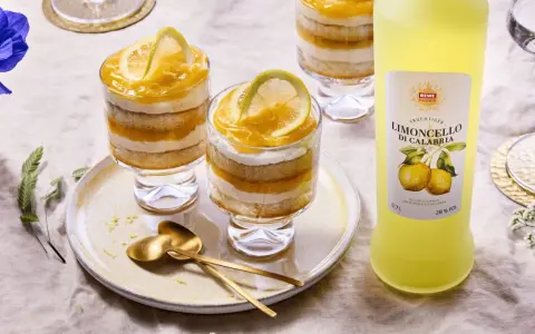 Erfrischendes Limoncello Tiramisu im Glas, geschichtet mit Biskuit und Zitronencreme, garniert mit Zitronenscheiben. Daneben eine Flasche Limoncello.