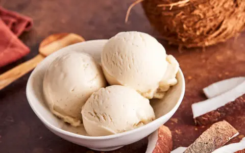 Schaufeln cremiges Kokoseis, perfekt zum Kokoseis selber machen mit Eismaschine. Daneben Kokosnussstücke.
