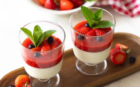 Zwei Gläser mit cremiger Panna cotta, garniert mit Erdbeersauce, frischen Erdbeeren, Blaubeeren und Minze.