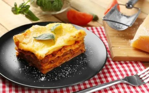 Ein Stück Lasagne mit Hackfleischsauce und cremiger Bechamelsoße, garniert mit Basilikum. Daneben ein Stück Parmesan und eine Gabel.