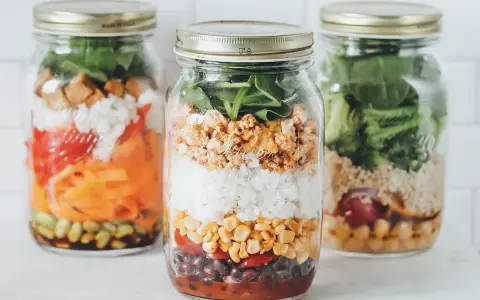 Drei Gläser mit geschichtetem Salat: Reis, Mais, Bohnen, Tomaten, Karotten und Spinat. Perfekt für unterwegs!