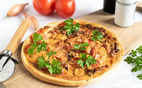 Flammkuchen mit Pfifferlingen, garniert mit Petersilie, auf einem Holzbrett. Tomaten und Schalotten im Hintergrund.
