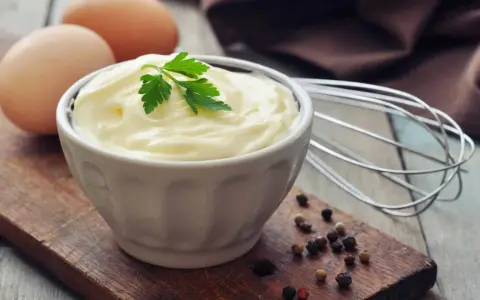 Cremige Mayonnaise in einer Schüssel, garniert mit Petersilie. Eier und ein Schneebesen deuten auf ein hausgemachtes mayonnaise rezept hin.