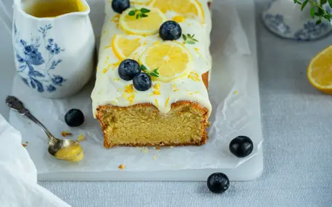 Ein saftiger zitronen schmand kuchen, dekoriert mit Zitronenscheiben, Blaubeeren und Zitrusschale. Daneben eine Kanne mit Zitronensauce.