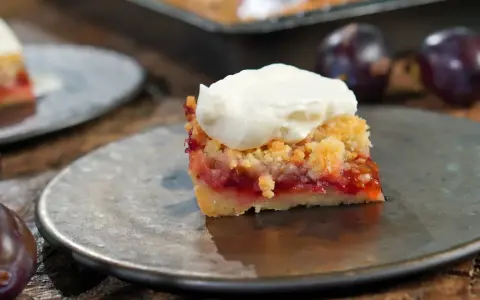 Ein Stück Zwetschgenkuchen mit Streuseln und Sahne auf einem Teller, daneben frische Zwetschgen.