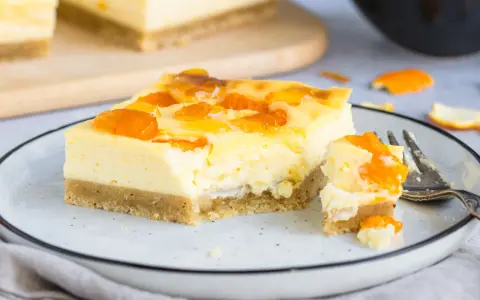 Ein Stück Mandarinen Schmand Kuchen Blech mit fruchtiger Glasur auf einem Teller, daneben ein Stück mit einer Gabel.