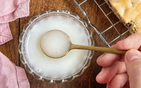 Ein Löffel mit zuckerguss ohne puderzucker in einer Schale. Daneben ein Stück Kuchen auf einem Gitter.