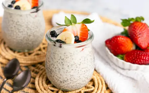 Zwei Gläser mit cremigem Chia Pudding, garniert mit Erdbeeren, Bananen und Blaubeeren. Daneben eine Schale voller frischer Erdbeeren.