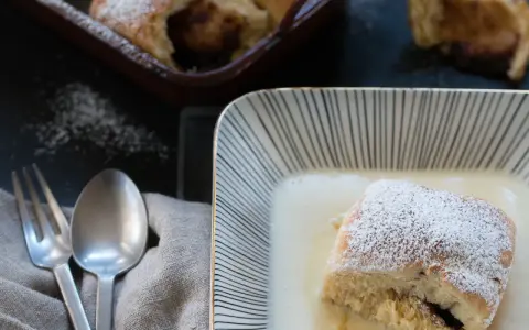 Buchteln original rezept: Ein Buchtel in Vanillesauce, bestäubt mit Puderzucker. Im Hintergrund weitere Buchteln im Ofen.