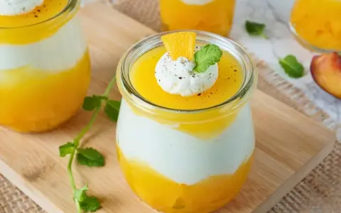 Erfrischendes Solero Dessert im Glas mit Mango-Fruchtsoße, weißer Creme und einem Klecks Sahne, garniert mit Minze und Orangenspalte.