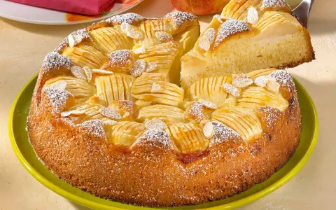 Ein saftiger, versunkener Apfelkuchen mit Mandelsplittern und Puderzucker bestreut, wird auf einem grünen Teller präsentiert.