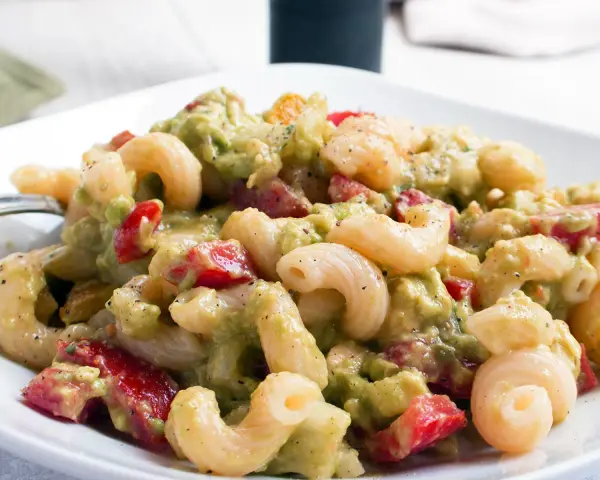 Ein cremiger nudelsalat mit avocado, Tomaten und Gewürzen auf einem weißen Teller.