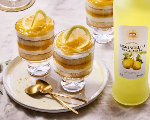 Erfrischendes Limoncello Tiramisu im Glas, geschichtet mit Biskuit und Zitronencreme, garniert mit Zitronenscheiben. Daneben eine Flasche Limoncello.