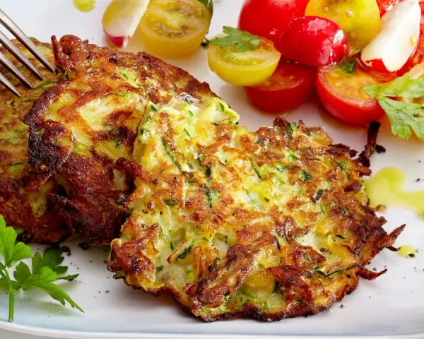 Goldbraune Zucchini-Puffer, knusprig gebraten, serviert mit einem bunten Tomatensalat und frischer Petersilie. Ein Genuss!